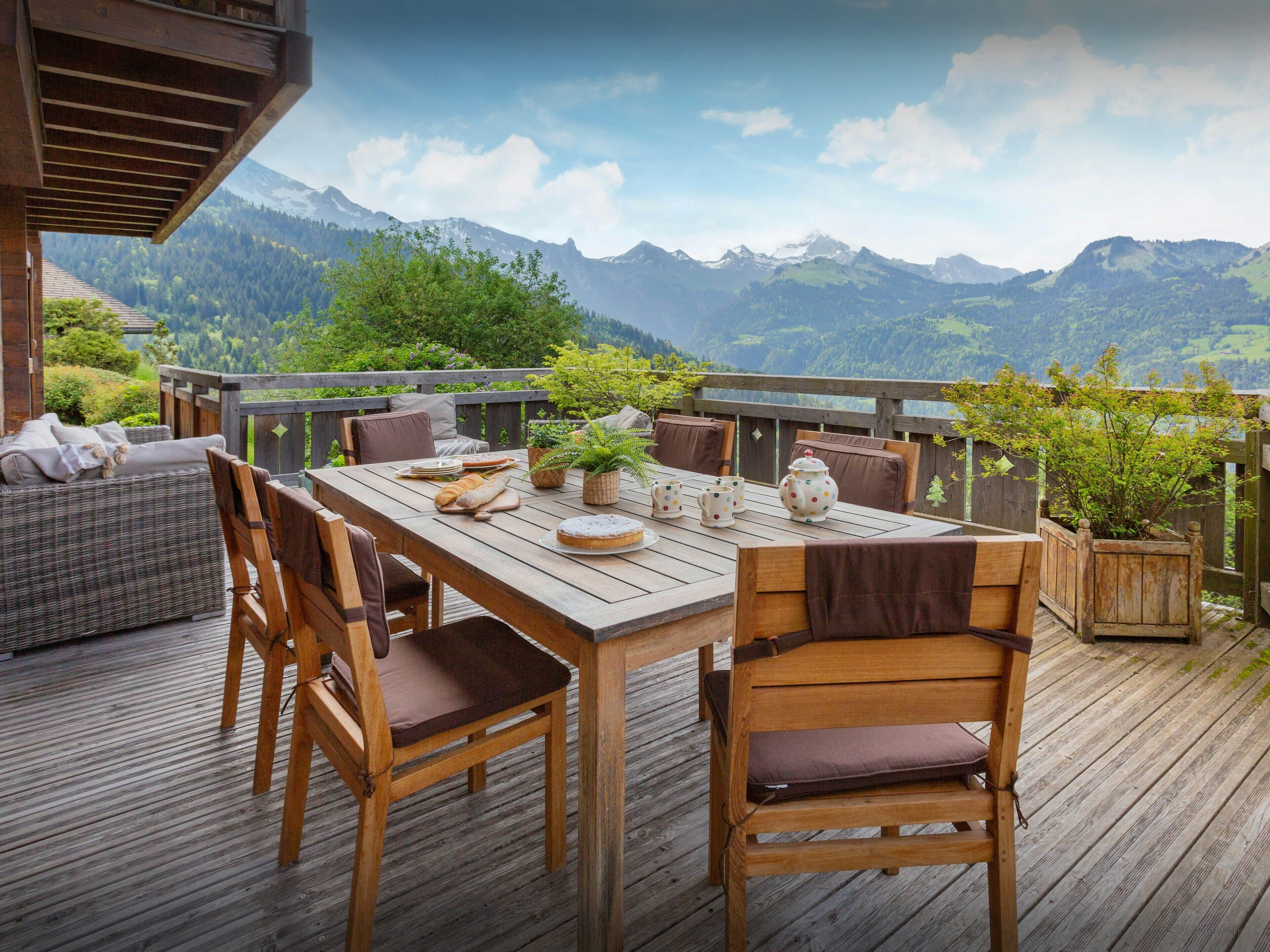 A wooden deck with a table set for dining, surrounded by chairs, overlooking a mountainous landscape.