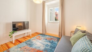 Un salon moderne avec un téléviseur à écran plat posé sur un meuble blanc, un tapis coloré, un canapé gris agrémenté de coussins, une petite table d’appoint et une fenêtre avec des rideaux.