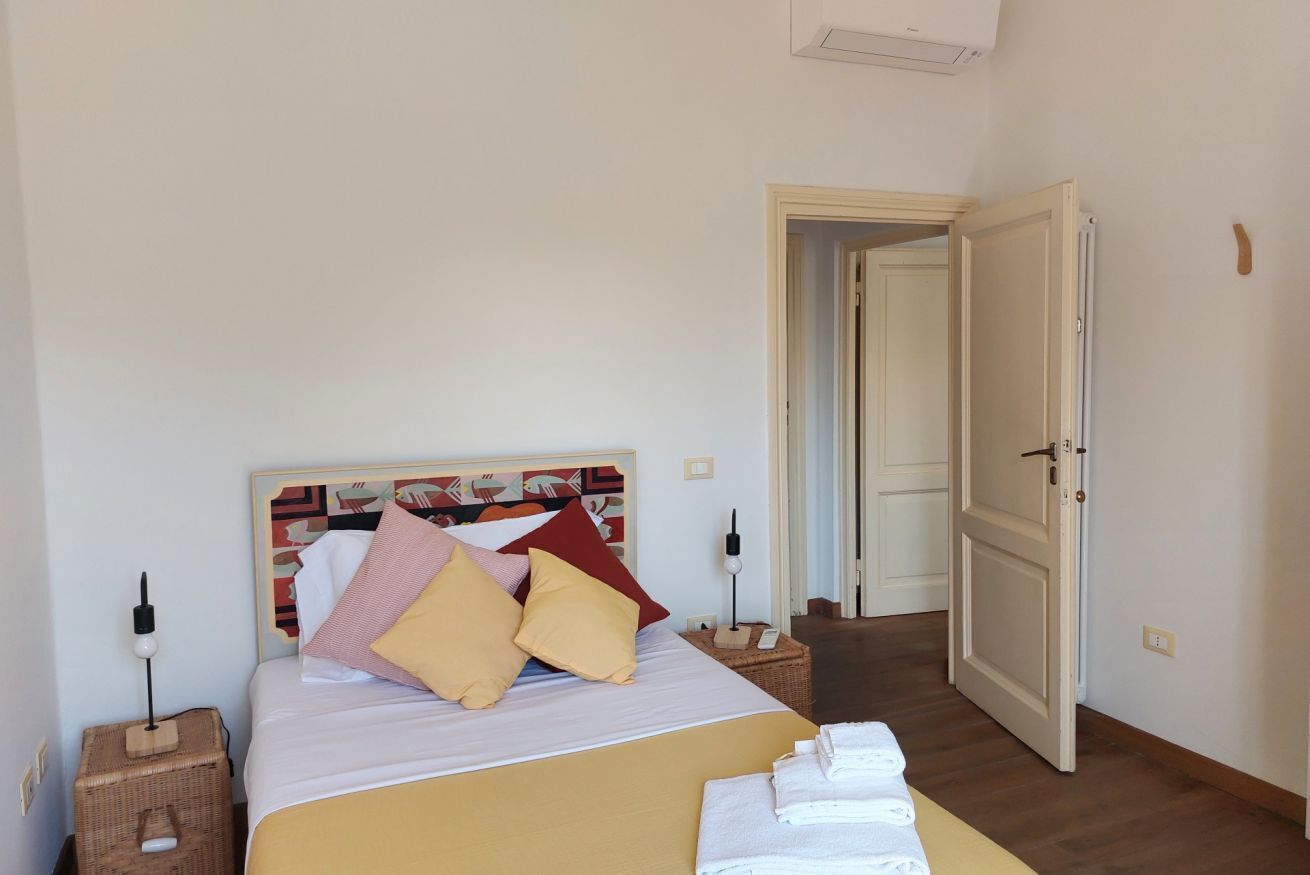 A neatly arranged bedroom with a bed, pillows, a bedside table, a lamp, and a door leading to another room.