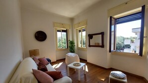 A living room with a white sofa, a small white table, and a white stool. There are two windows with blinds, a potted plant, and a mirror on the wall. - Beautiful penthouse with loggia and air conditioning, 100 meters from the sea. (Viareggio)