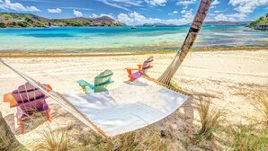 Une plage avec un hamac, des chaises colorées et une vue dégagée sur la mer et les collines lointaines.