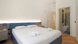 Moderni hotellihuone, jossa on suuri sänky, yöpöydät, yölamppu ja avoimesta ovesta näkyvä kylpyhuone.