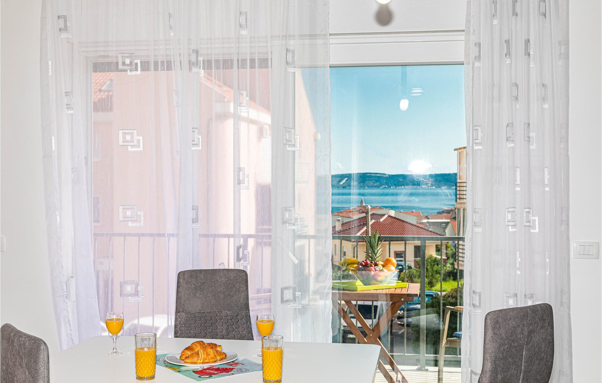 A dining area with a table set for breakfast, including croissants and orange juice, and a view of a coastal town through large windows.