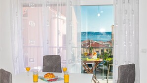 Ein Essbereich mit einem für das Frühstück gedeckten Tisch, darunter Croissants und Orangensaft, und Blick auf eine Küstenstadt durch große Fenster.