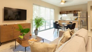 A modern living room with a sofa, a coffee table, and a TV. There is a dining area with chairs and a table. A large window provides natural light. - Caseiba Peaceful Stays 7 (Mérida Yucatan)