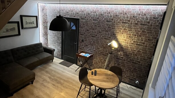 Ein gemütliches Loft mit freiliegenden Backsteinwänden, einem Holzesstisch mit Stühlen, einer dunklen Couch und einer schwarzen Pendelleuchte.