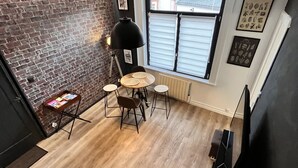 Ein gemütliches Zimmer mit Backsteinwand, Holzboden, einem runden Tisch mit Stühlen, einer schwarzen Lampe und einem Fenster mit Jalousien.