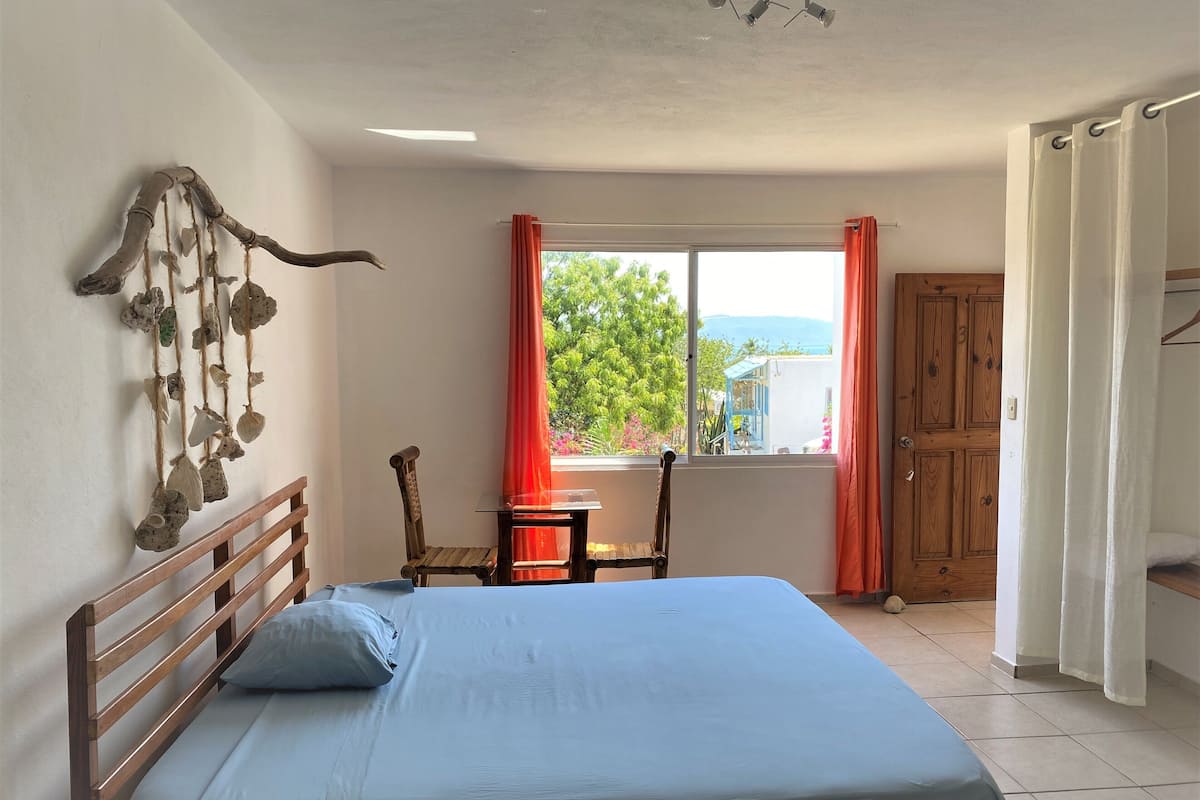 A bedroom with a bed, a wooden headboard, a small table with chairs, a window with curtains, and a door.
