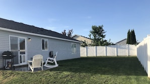 En trädgård med en gräsmatta, två Adirondack-stolar, en grill och ett vitt staket.