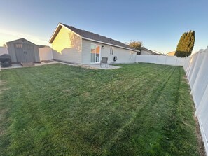 A residential backyard with a house, a shed, a lawn, and a white fence. - Perfect Space for a Comfortable Stay (Pasco)