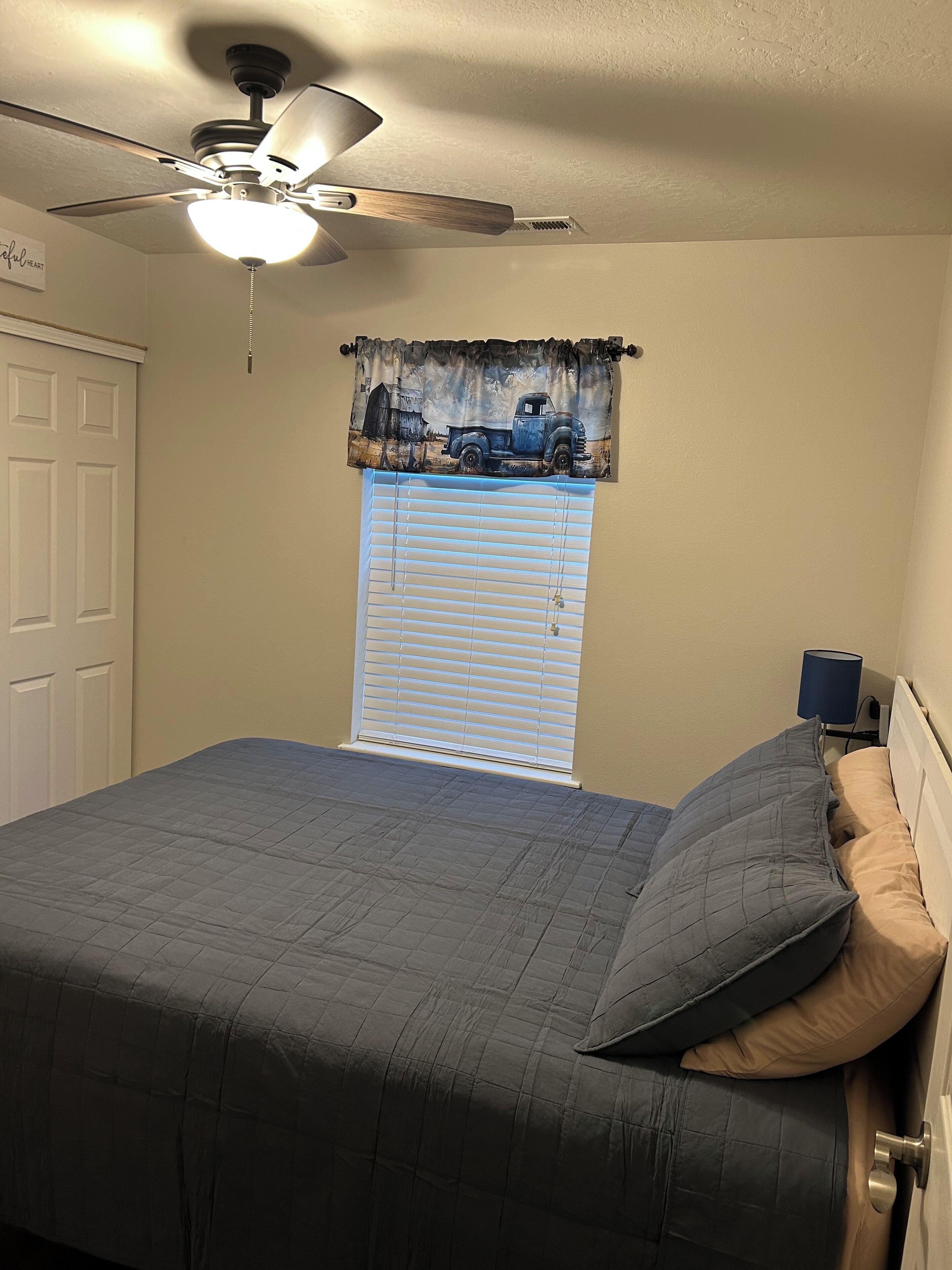 Une chambre à coucher comprenant un lit, un ventilateur de plafond, une fenêtre avec des stores et une table de chevet avec une lampe.