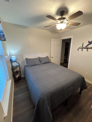 Un dormitorio con cama, ventilador de techo, mesita de noche con lámpara y puerta que da a otra habitación.