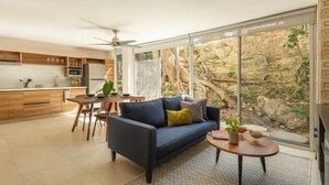 Un salon moderne avec un canapé bleu foncé, une table basse ronde et un coin repas avec des chaises en bois. Une grande fenêtre offre une vue sur un espace extérieur rocheux.