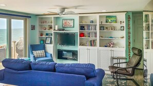 Un soggiorno con un divano blu, una poltrona, una libreria e vista sull'oceano tramite una porta scorrevole.