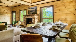 Un intérieur en bois avec une table à manger dressée pour six personnes, une cheminée et de grandes fenêtres offrant une vue sur les montagnes.
