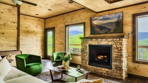 L’intérieur chaleureux d’un chalet avec une cheminée en pierre, des murs en bois et de grandes fenêtres offrant une vue panoramique.