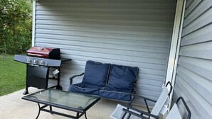 Eine Terrasse mit einem Glastisch, zwei blauen Sitzgelegenheiten, einem Grill und einem kleinen Tisch mit einem roten Gegenstand darauf.