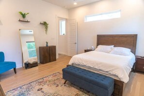 A bedroom with a large bed, a blue bench, a wooden dresser, a mirror, and a small plant.