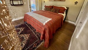 Un dormitorio con piso de madera, una cama con manta roja, un tapete estampado, un espejo adosado a la pared y una lámpara en el techo.