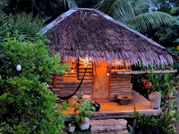 A rustic thatched-roof hut with a wooden door, a small porch, and potted plants around it.