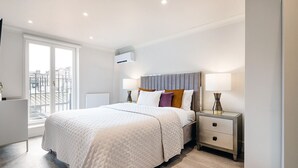 Una camera da letto moderna con un letto grande, un comodino con due lampade e vista sulla città attraverso un'ampia finestra.