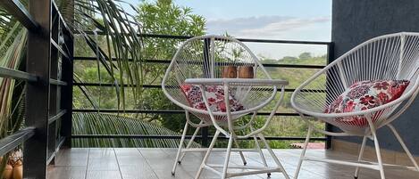 Ein Balkon mit zwei weißen Stühlen aus Draht, einem Kachelboden und Blick auf Grünpflanzen und Himmel.
