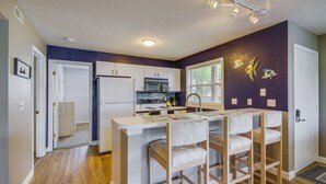 A compact kitchen with a microwave, stove, and refrigerator. There is a small island with bar stools and a window with decorative fish art. - Osage Beach Condo w/ Private Deck & Lake Views! (Osage Beach)