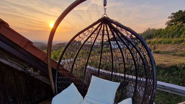 A hanging chair with cushions on a wooden deck, overlooking a landscape with trees and a sunset. - Cottage with whirlpool and sauna (Stara Gora)