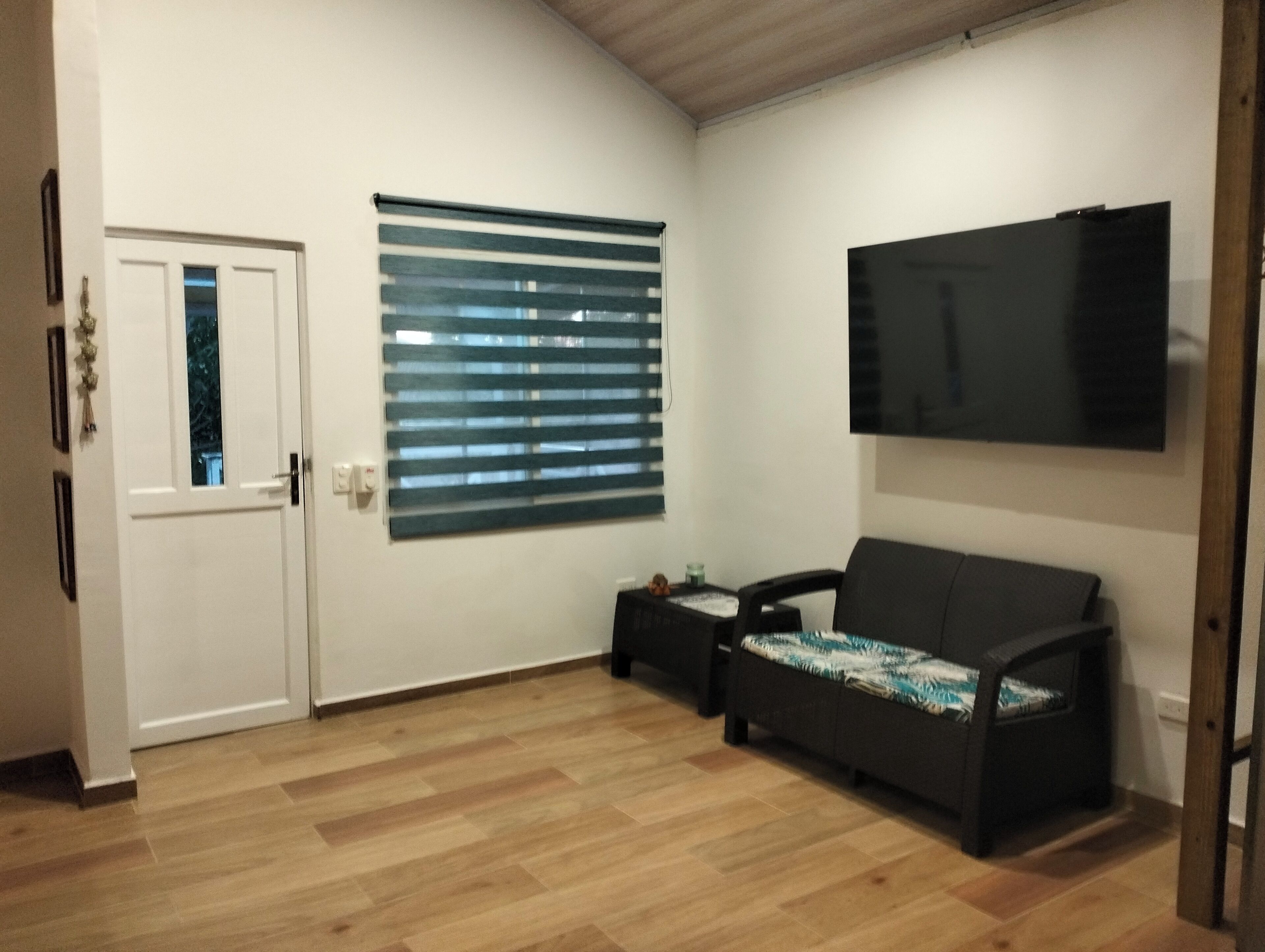 A room with a white door, a window with blinds, a black sofa with a patterned cover, a small table with a candle, and a flat-screen TV mounted on the wall.