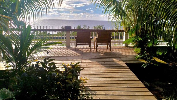 Una terraza de madera con dos sillas de madera y una barandilla, rodeada de exuberante vegetación tropical.
