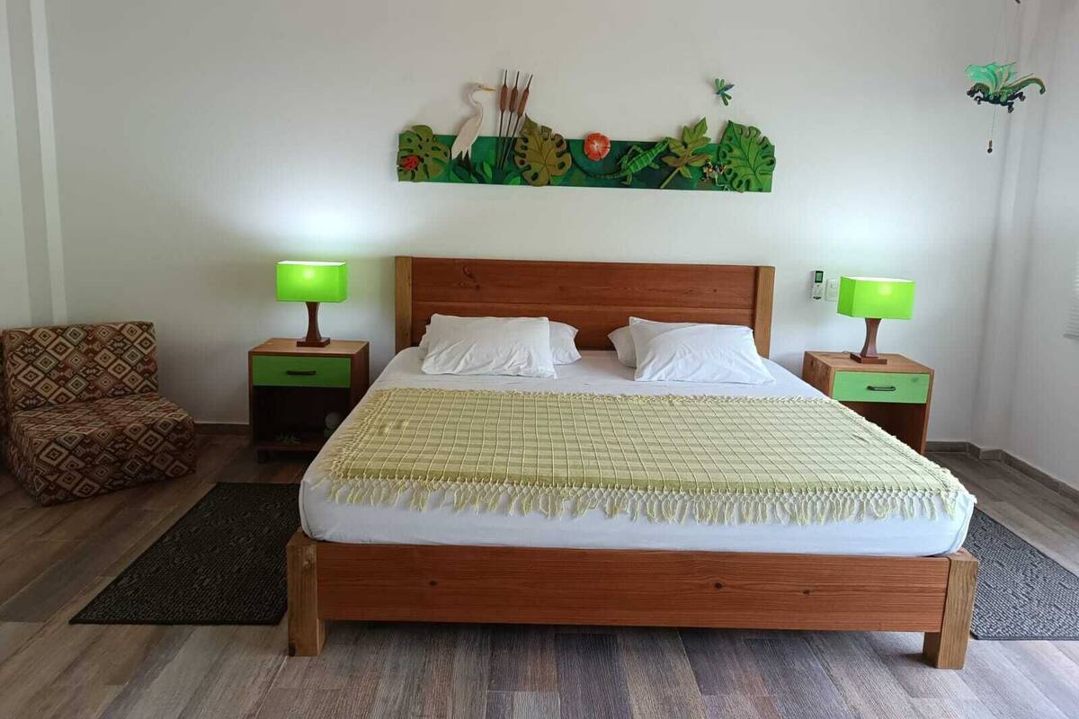Ein ordentlich eingerichtetes Schlafzimmer mit einem hölzernen Bett, zwei Nachttischen mit Lampen, einem gemusterten Sessel und einem Wandbild.