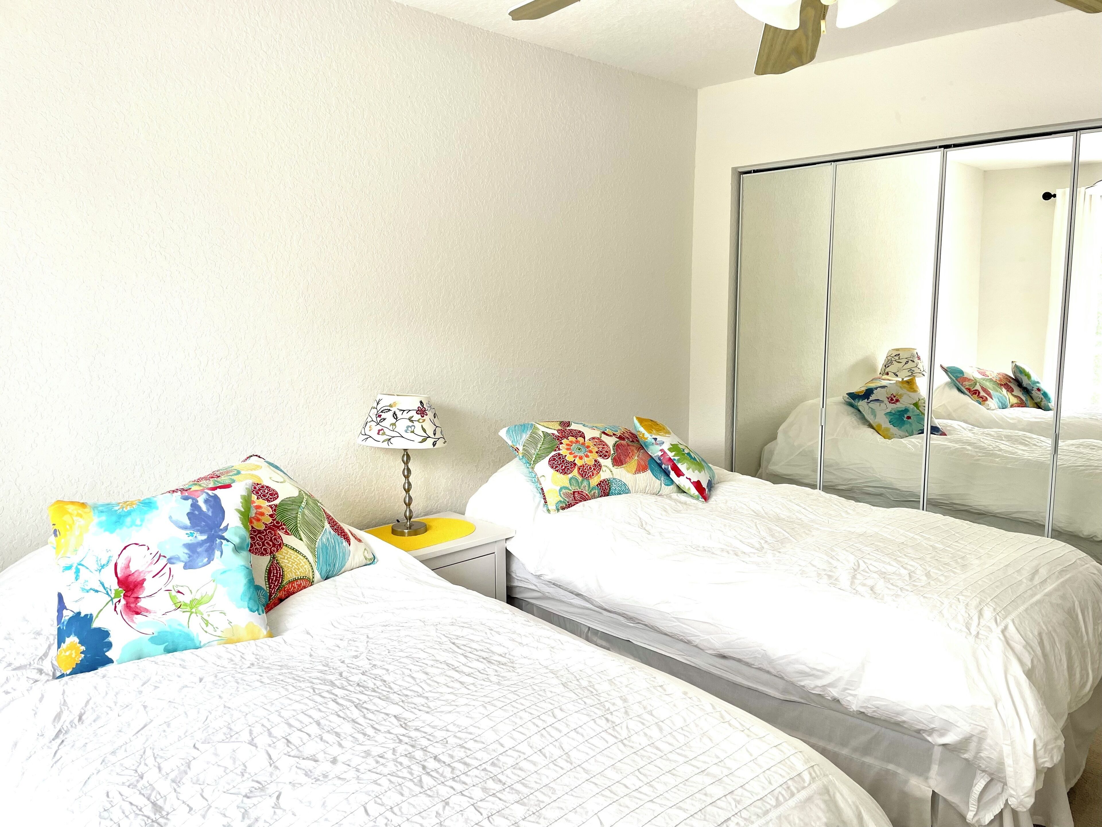 Een hotelkamer met twee bedden, een nachtkastje met een lamp, een spiegel en kussens met een bloemenprint.