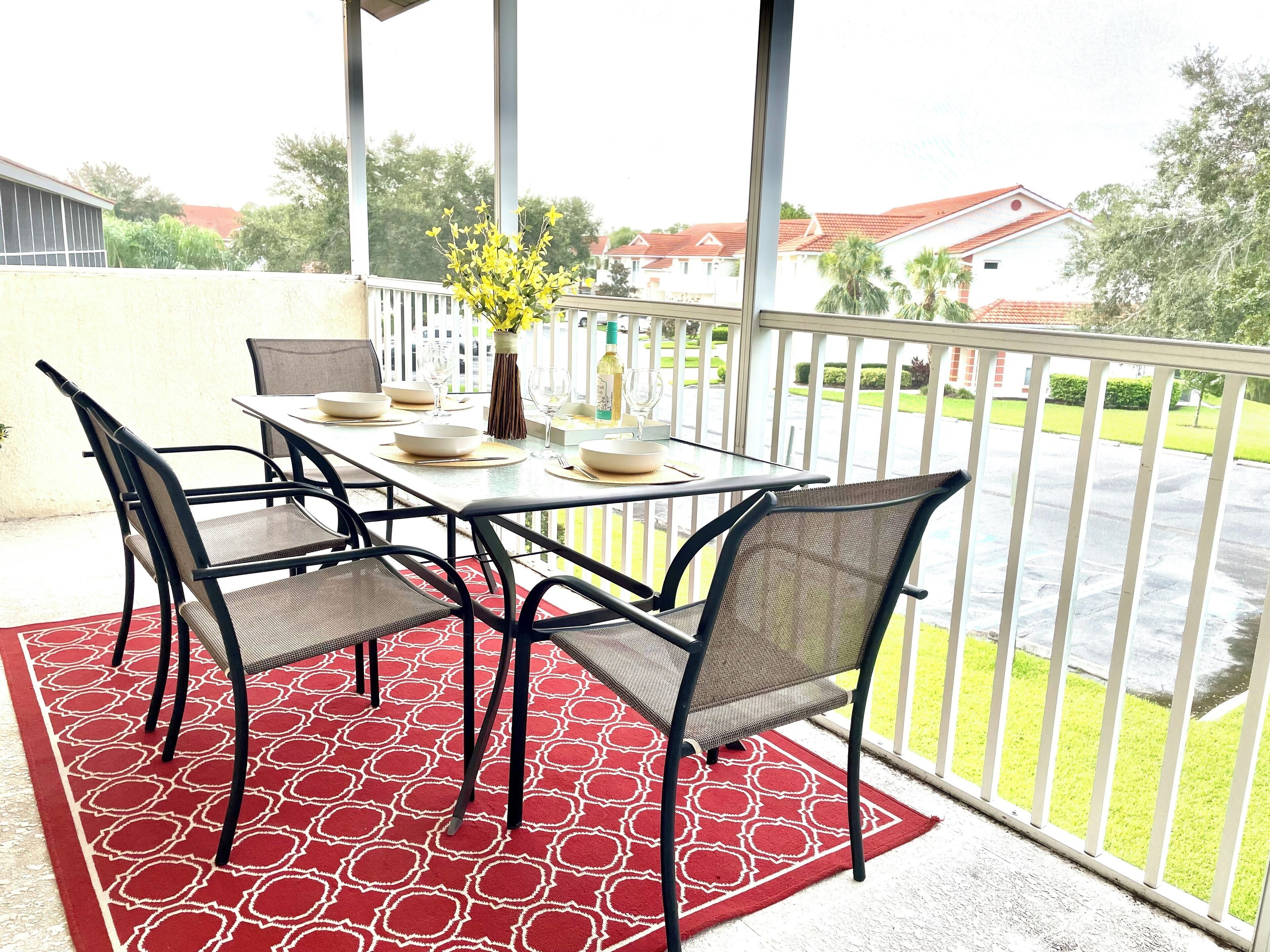 A covered patio with a table set for four, surrounded by chairs, a red patterned rug, and a vase with yellow flowers.