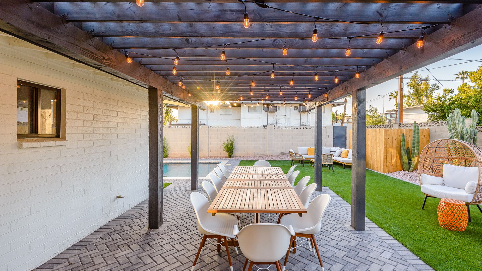 Un patio al aire libre con mesa y sillas de madera, una pérgola con luces colgantes y una piscina al fondo.