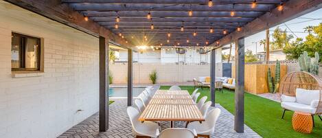 Un patio al aire libre con mesa y sillas de madera, una pérgola con luces colgantes y una piscina al fondo.