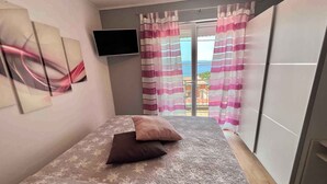 Une chambre à coucher avec un lit, un téléviseur à écran plat et une vue sur la mer, visible à travers les rideaux.