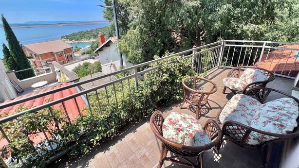 Ein Balkon mit Korbstühlen und einem Tisch, mit Blick auf ein Gewässer und Gebäude.