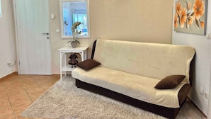 Ein Wohnzimmer mit einem beigen Sofa, einem kleinen weißen Beistelltisch mit einer Topfpflanze, einem Spiegel und einem Blumenbild an der Wand.