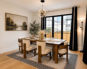 A dining room with a wooden table, six chairs, a vase with dried plants, a painting, and a large window with curtains.