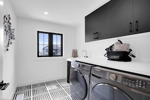 A modern laundry room with a washing machine, dryer, and a black laundry basket on the counter. - Brand New Modern Luxury Villa (London)