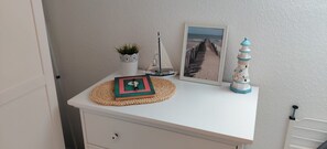 A white bedside table with a framed picture of a beach, a small potted plant, a decorative lighthouse, and a book.