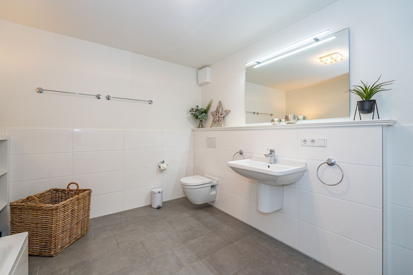 Ein modernes Badezimmer mit Waschbecken, Toilette und einem großen Spiegel. Auf dem Boden steht ein Korbwäscher und ein kleiner Mülleimer.