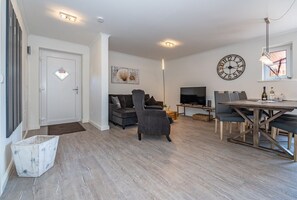 Een moderne woonkamer met een grijze bank, een televisie en een eethoek met een houten tafel en stoelen.