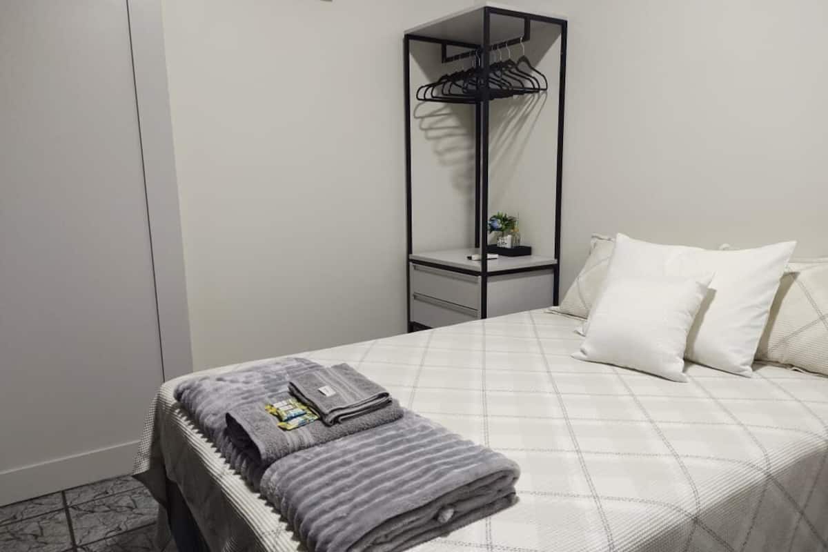 A neatly made bed with a grey blanket and a folded towel, a black metal wardrobe, a small bedside table with a plant, and white pillows.