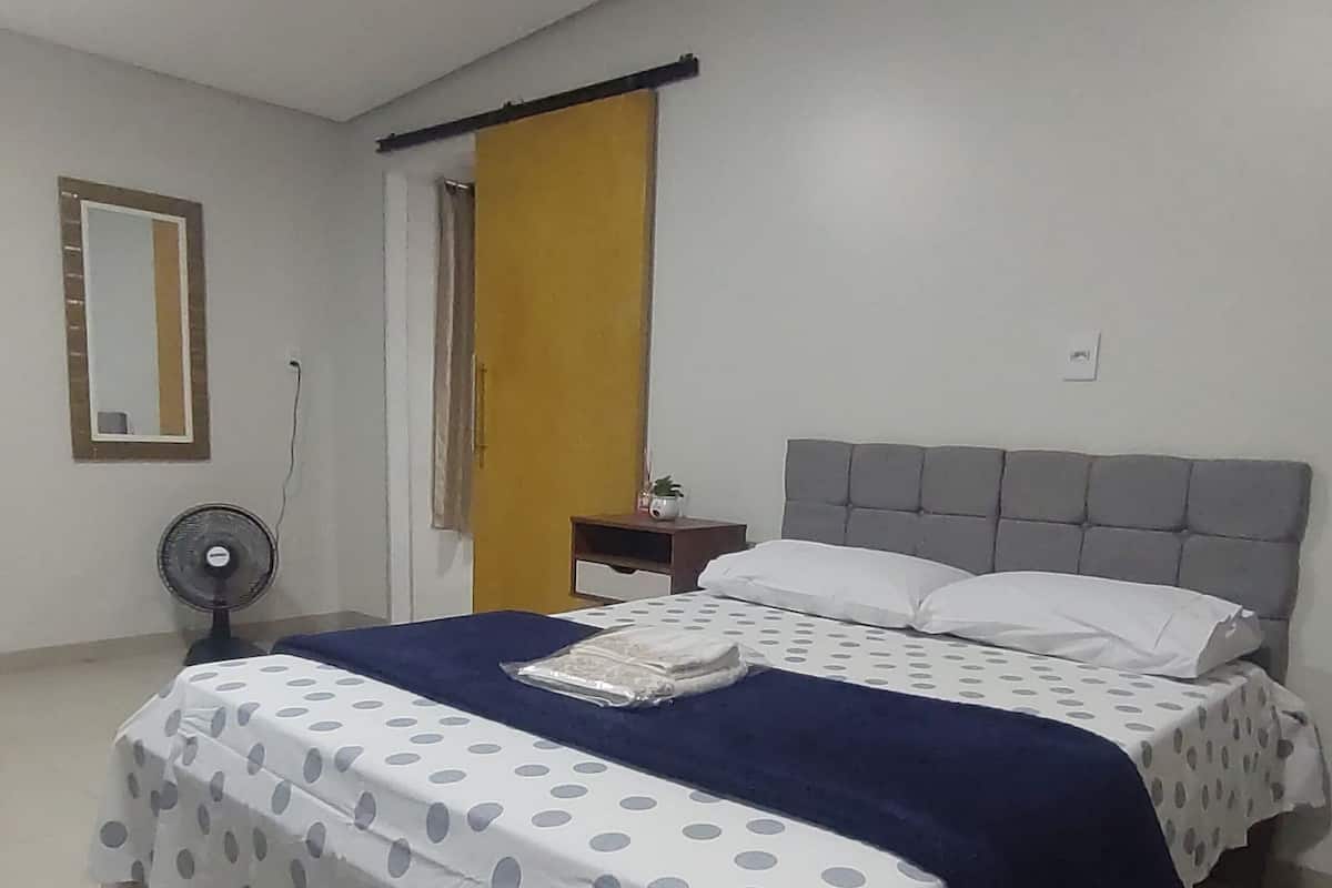 A bedroom with a bed, a bedside table, a nightstand, a mirror, a fan, and a window with curtains.