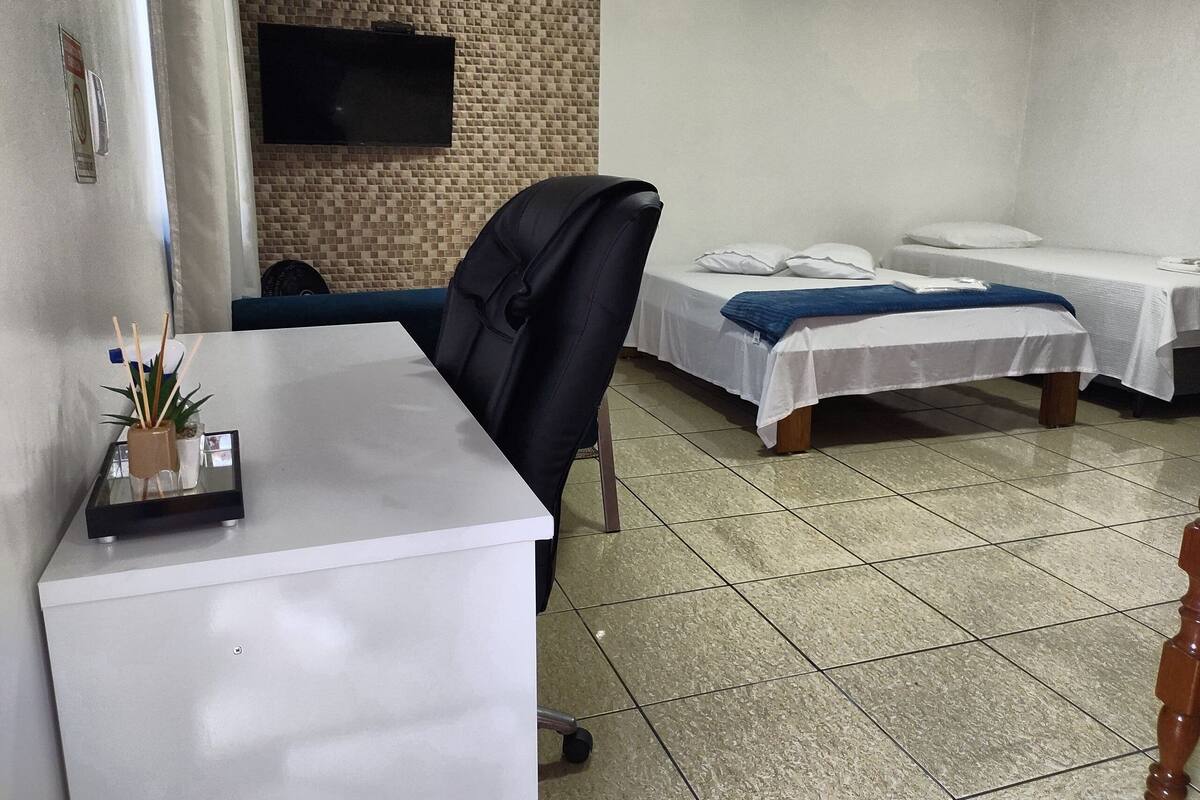 A room with a desk, a chair, two beds, a TV on the wall, and a tiled floor.