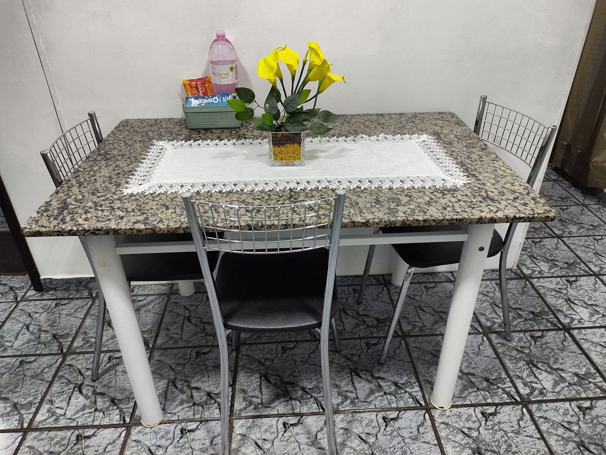Ein Esstisch mit weißen Tischdecken, vier Stühlen, einer Vase mit gelben Blumen, einer Wasserflasche und einer Tüte Snacks.