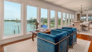 Ein geräumiges Zimmer mit großen Fenstern und Blick auf ein Gewässer, ausgestattet mit blauen Polsterstühlen, einem Essbereich und einem Kronleuchter.