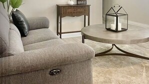 En stue med en grå sofa, et rundt sofabord, et spejl på væggen, et lille træsidebord med dekorative genstande, og et moderne kunstværk.