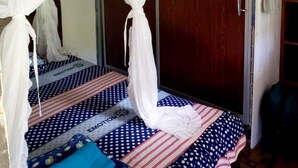 Quarto com cama coberta por um dossel branco, porta de madeira e prateleira pequena com toalhas.
